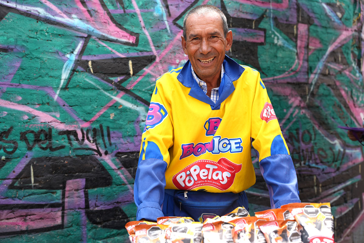Un hombre posa sonriente frente a una pared con grafitis llenos de color. Viste uniforme de Bon Ice y PoPetas, mientras ofrece sus productos en la vía pública